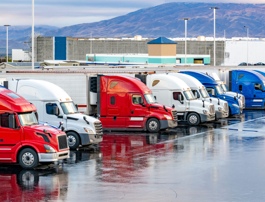 Different make big rigs semi trucks tractors with loaded semi trailers standing in the row on truck stop parking lot at early morning waiting for the route continuation time according to the log book