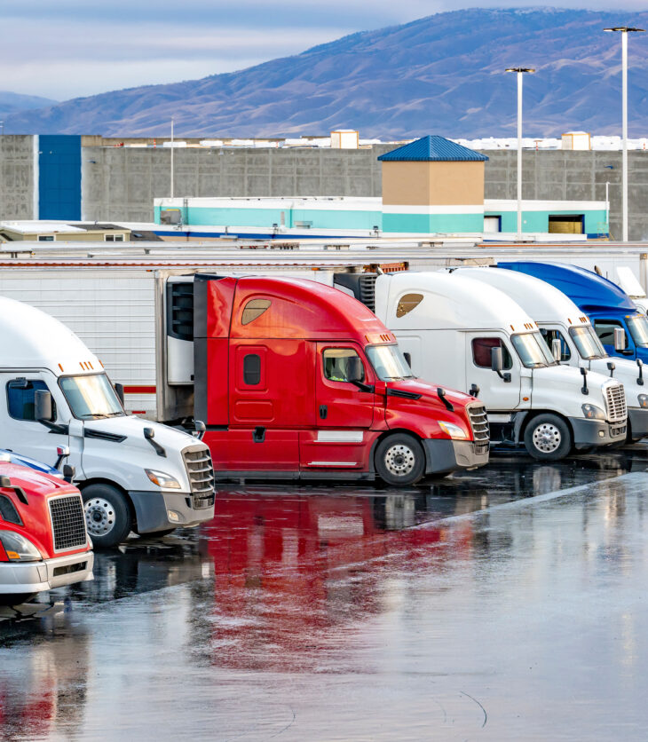 Different make big rigs semi trucks tractors with loaded semi trailers standing in the row on truck stop parking lot at early morning waiting for the route continuation time according to the log book